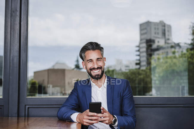Homme d'affaires souriant avec écouteurs intra-auriculaires et téléphone portable appuyé sur la fenêtre — Photo de stock