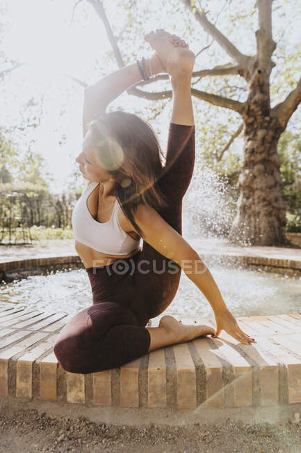 Sportswoman practicing compass pose while sitting by fountain at park — Stock Photo