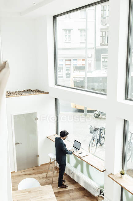 Männlicher Unternehmer benutzte Laptop, während er im Büro am Glasfenster stand — Stockfoto