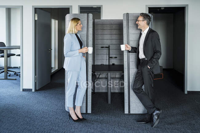 Uomo d'affari sorridente e donna d'affari con tazza di caffè parlare durante la pausa mentre in piedi vicino al cubicolo dell'ufficio — Foto stock