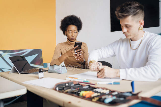 Молодая женщина, использующая смартфон, пока мужчина работает за столом в студии — стоковое фото