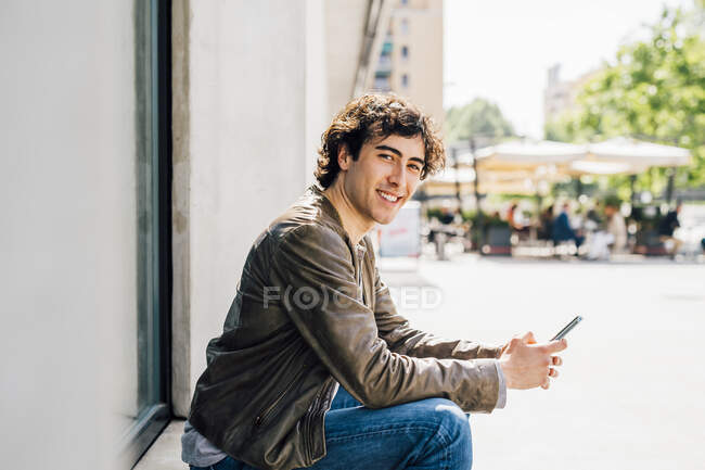 Усміхнений чоловік тримає мобільний телефон, сидячи біля дверей. — Stock Photo