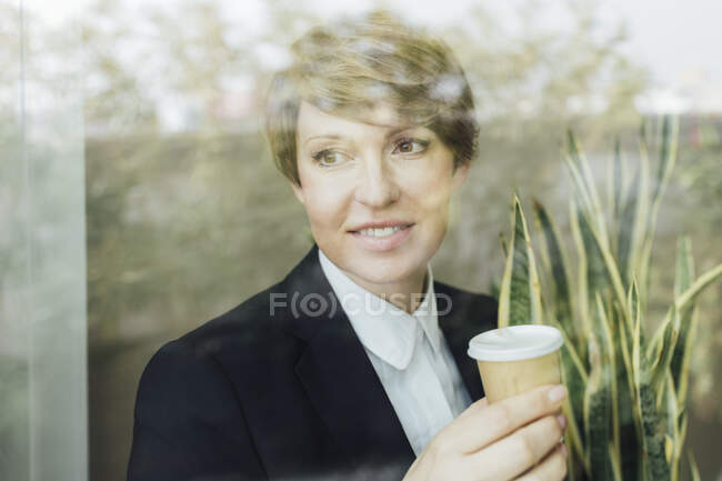 Lächelnde Unternehmerinnen, die wegschauen, während sie Einweg-Kaffeetassen aus dem Glasfenster halten — Stockfoto
