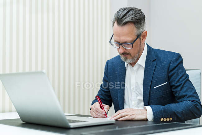 Geschäftsmann mit Laptop schreibt während der Arbeit im Büro in Notizblock — Stockfoto