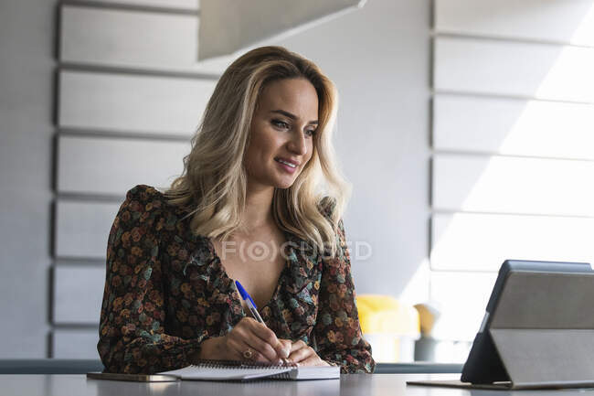 Female entrepreneur looking at digital tablet writing on diary at office — Stock Photo