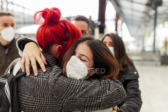 Mujeres amigas con máscaras que se abrazan en la estación de tren durante COVID-19 - foto de stock
