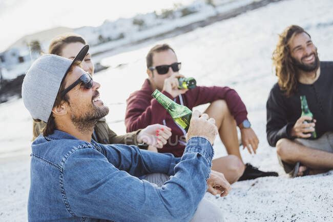 Homme souriant prenant un verre tout en étant assis avec des amis à la plage — Photo de stock