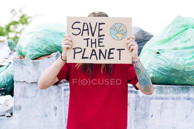 Hippie man covering face with save the planet cardboard near garbage dump — Stock Photo