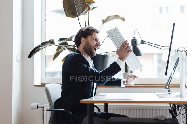 Angry businessman holding computer keyboard while sitting by desk — Stock Photo