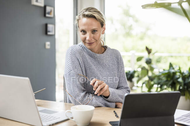 Empresaria sosteniendo anteojos mientras está sentada junto a la computadora portátil y la tableta digital en casa - foto de stock