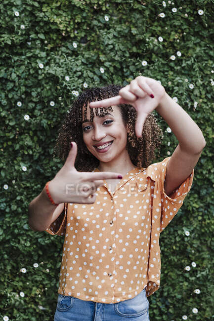 Sorridente giovane donna facendo cornice dito davanti alla siepe — Foto stock