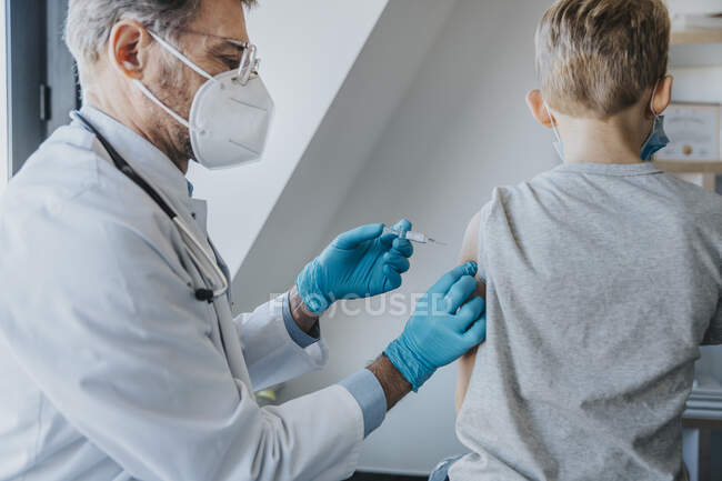 Доктор носить захисну маску обличчя, даючи COVID-19 вакцину хлопчику, стоячи в клініці — стокове фото