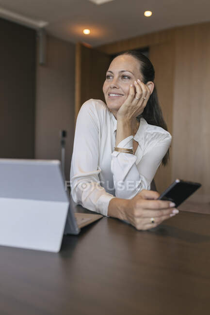 Усмішка дбайлива жінка-професіонал, яка тримає смартфон за столом у кабінеті. — стокове фото