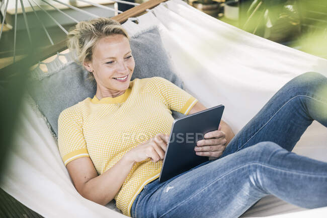 Усміхнена жінка користується цифровим планшетом, відпочиваючи в гамаку. — стокове фото