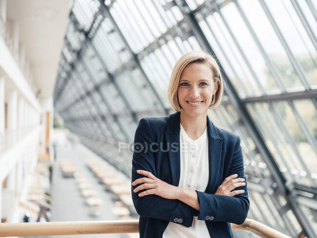Mujer de negocios sonriente con los brazos cruzados apoyados en barandilla en la oficina - foto de stock