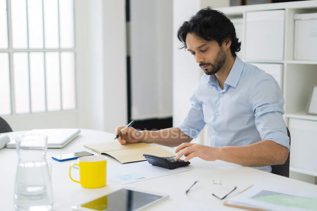 Jeune entrepreneur utilisant une calculatrice pendant qu'il travaille au bureau — Photo de stock