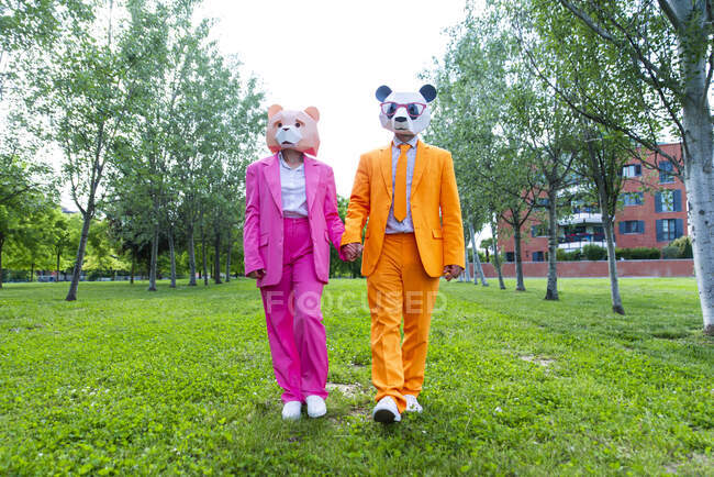 Coppia adulta che indossa abiti vibranti e maschere animali che si tengono per mano mentre camminano insieme nel parco verde — Foto stock