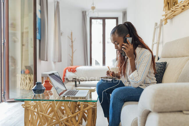 Mulher falando ao telefone enquanto escrevia na sala de estar em casa — Fotografia de Stock