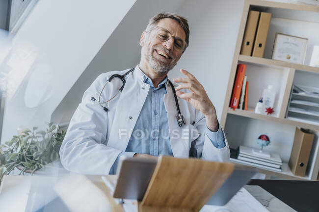 Hombre trabajador de la salud hablando en videollamada con el paciente a través de tableta digital mientras está sentado en el consultorio del médico - foto de stock