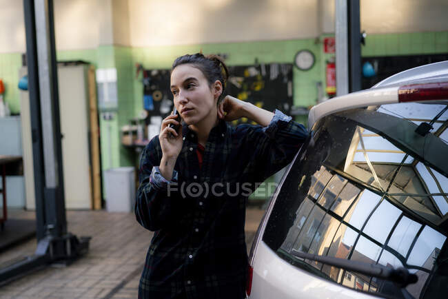 Feminino mecânico olhando afastado falando no telefone inteligente na oficina de reparação — Fotografia de Stock