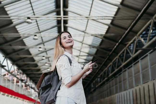 Cheerful young female passenger wearing backpack looking away at railroad station — Stock Photo