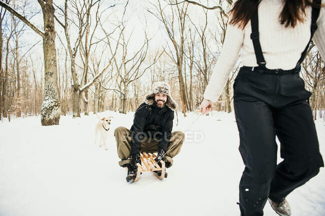 Жінка тягне чоловіка на дерев'яній сані з собакою позаду у лісі. — стокове фото