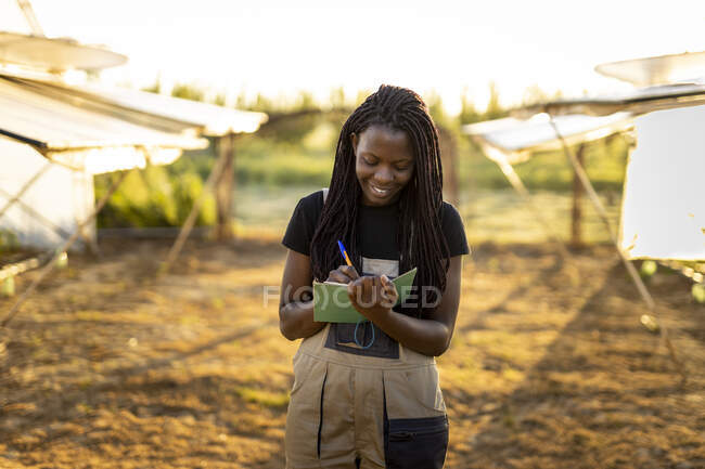 Lächelnde Bäuerin mit langen Haaren schreibt auf Notizblock, während sie auf dem Hof steht — Stockfoto