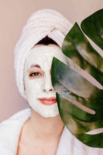 Junge Frau mit Schönheitsgesichtsmaske, die Auge mit Monstera-Blatt bedeckt — Stockfoto