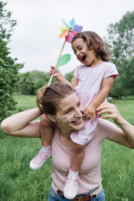 Laughing daughter holding pinwheel while sitting on mother's shoulder at park — Stock Photo