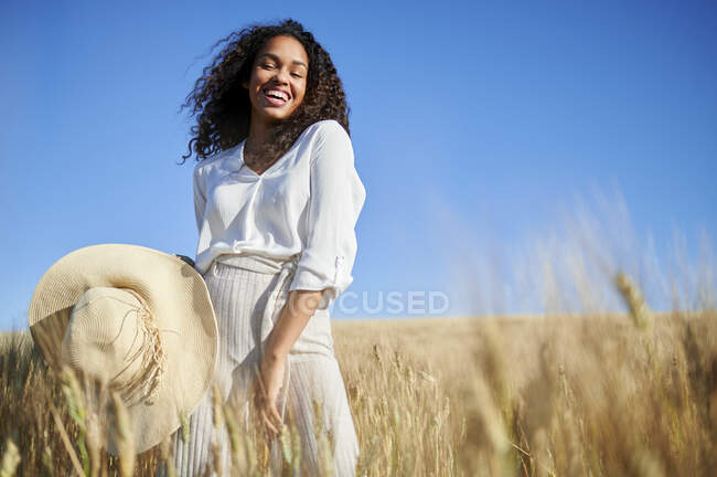 Весела кучерява жінка з капелюхом, що стоїть на пшеничному полі в сонячний день — стокове фото
