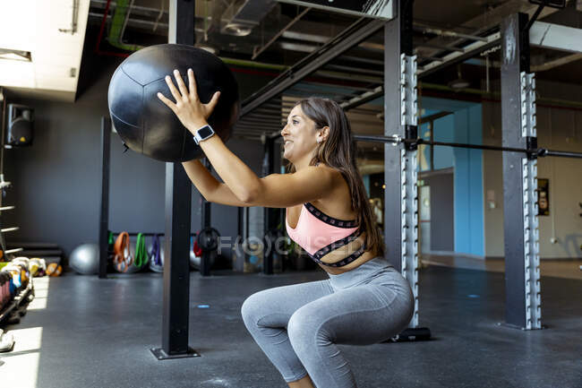 Smiling atleta feminina agachado durante o exercício com bola de fitness no ginásio — Fotografia de Stock