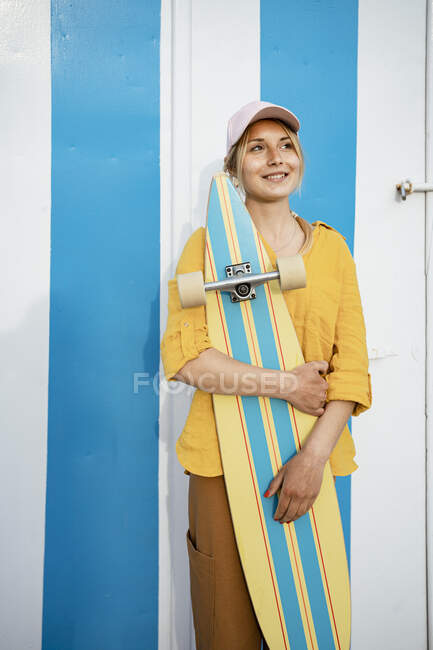 Sonriente joven mujer sosteniendo longboard día soñando mientras mira hacia otro lado - foto de stock