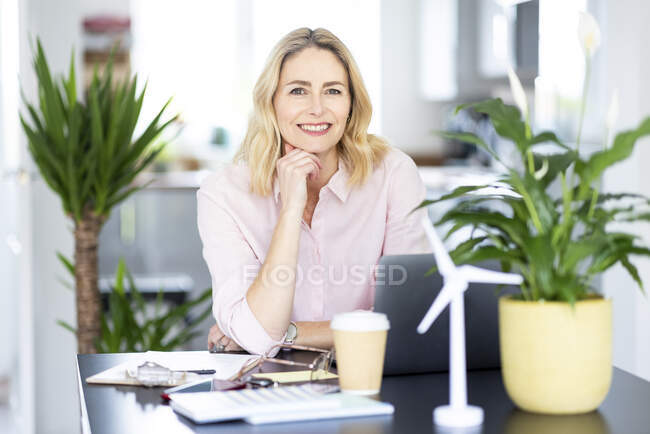 Donna d'affari sorridente con la mano sul mento seduta alla scrivania in ufficio — Foto stock