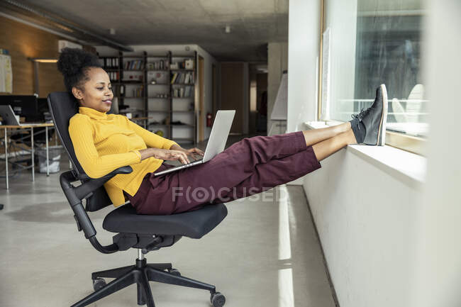 Profissional feminino usando laptop enquanto se senta com os pés para cima na cadeira no escritório — Fotografia de Stock