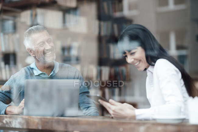 Male and female owners with laptop and mobile phone smiling by cafe window — Stock Photo