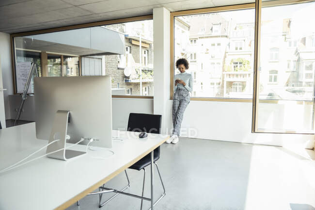 Professionista femminile utilizzando lo smartphone mentre si appoggia alla finestra in ufficio — Foto stock