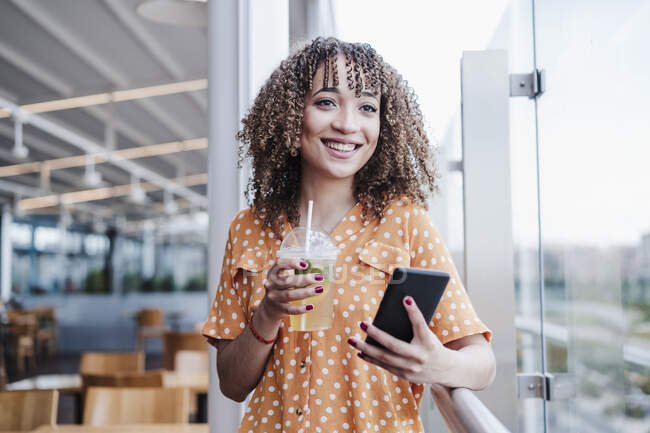 Giovane donna sorridente con il telefono cellulare che distoglie lo sguardo mentre ha tè freddo nel balcone al caffè — Foto stock