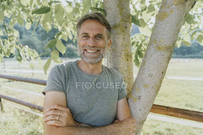 Lächelnder älterer Mann mit verschränkten Armen, an Baum gelehnt — Stockfoto