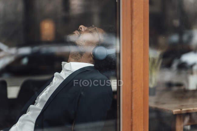 Männlicher Profi mit geschlossenen Augen hört Musik durch Kopfhörer im Café durch Glas gesehen — Stockfoto