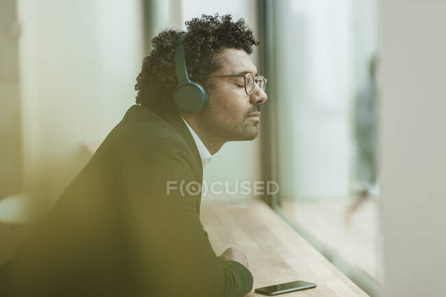 Männlicher Profi mit geschlossenen Augen hört Musik über Kopfhörer, während er sich im Büro auf den Schreibtisch lehnt — Stockfoto
