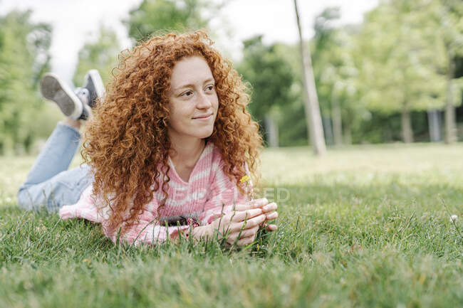 Donna sorridente con i capelli ricci sdraiati sull'erba al parco guardando altrove — Foto stock