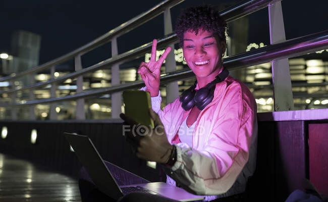 Весела жінка, що показує знак перемоги вночі під час відвідин смарт-телефону — стокове фото