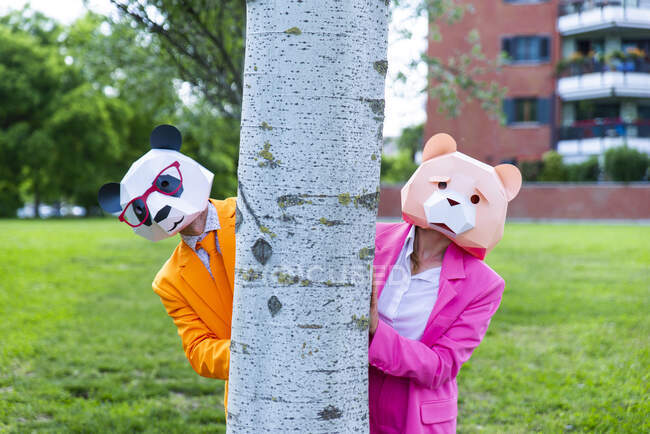 Man and woman wearing vibrant suits and animal masks hiding together behind birch tree — Stock Photo