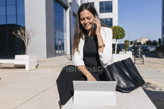 Усміхаючись, жінка - підприємець розмовляє по телефону, користуючись цифровим планшетом у сонячний день. — стокове фото