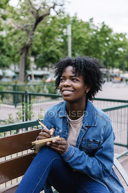 Sonriente mujer pensativa mirando hacia arriba mientras escribe en bloc de notas - foto de stock