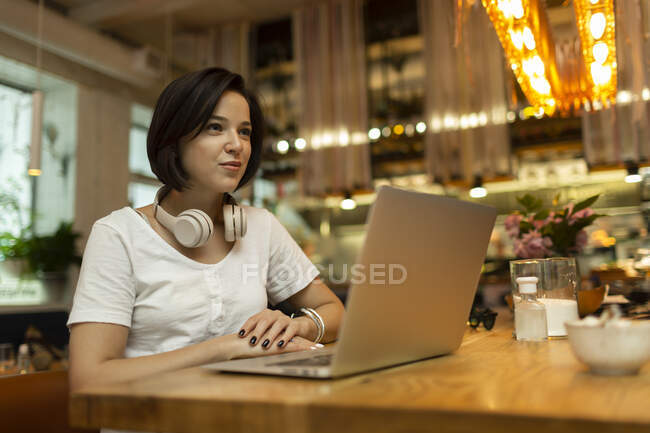 Усміхнена молода жінка-фрілансер сидить з ноутбуком в ресторані, дивлячись у далечінь. — стокове фото