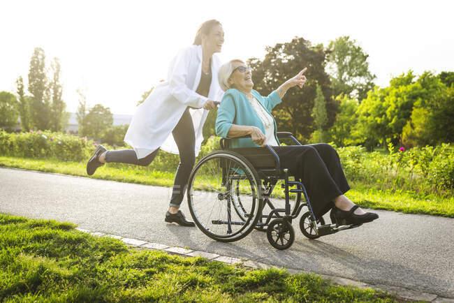 Femme âgée joyeuse pointant tandis que le gardien roulant fauteuil roulant dans le parc — Photo de stock