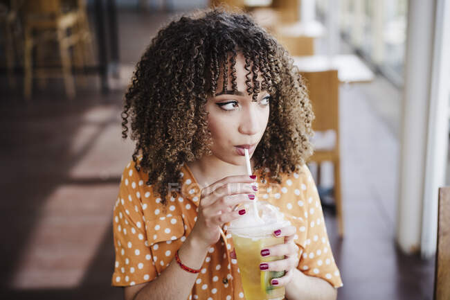 Молодая женщина с вьющимися волосами смотрит в сторону, когда пьет чай со льдом в кафе — стоковое фото