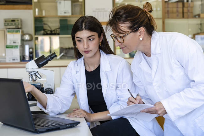 Científica escribiendo en documento mientras discute con su colega sobre el portátil - foto de stock