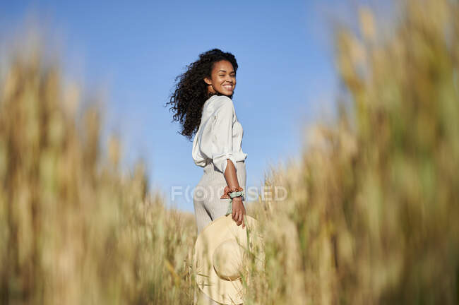 Усміхнена кучерява жінка з капелюхом, що стоїть на пшеничному полі в сонячний день — стокове фото